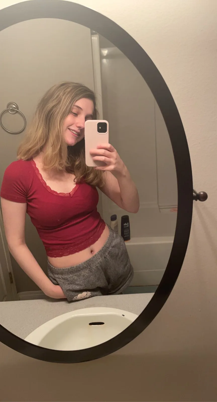 A young woman with blonde hair takes a mirror selfie in a bathroom, wearing a red crop top and grey shorts, smiling while…