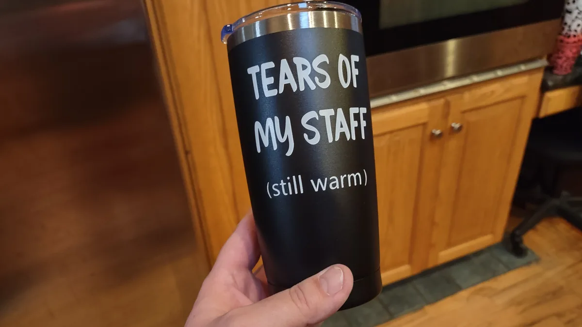 A hand holds a black insulated tumbler with white text reading 'TEARS OF MY STAFF (still warm)' against a background of…