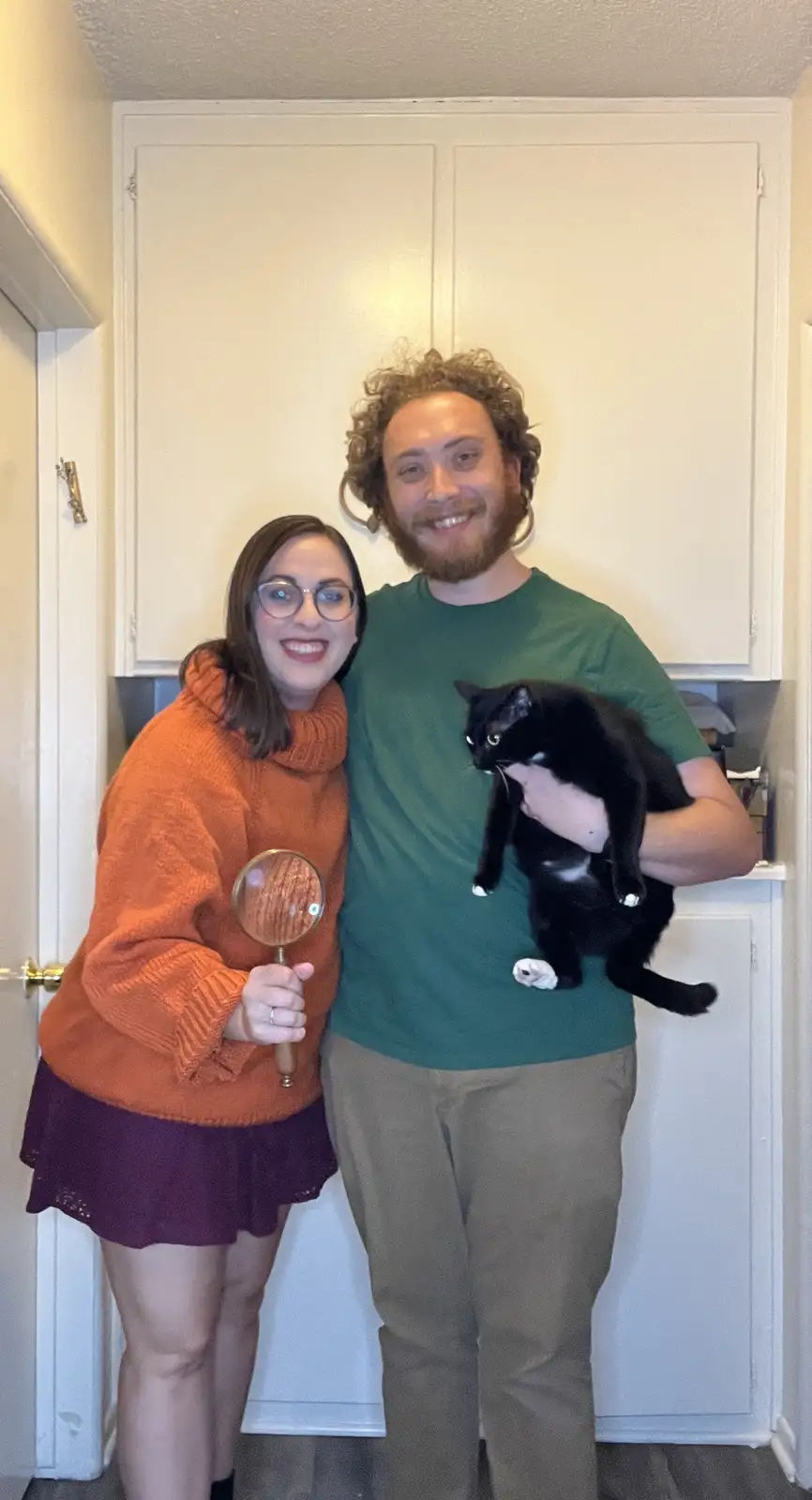 A man with curly hair and a beard, wearing a green t-shirt and holding a black cat, stands next to a woman in an orange…