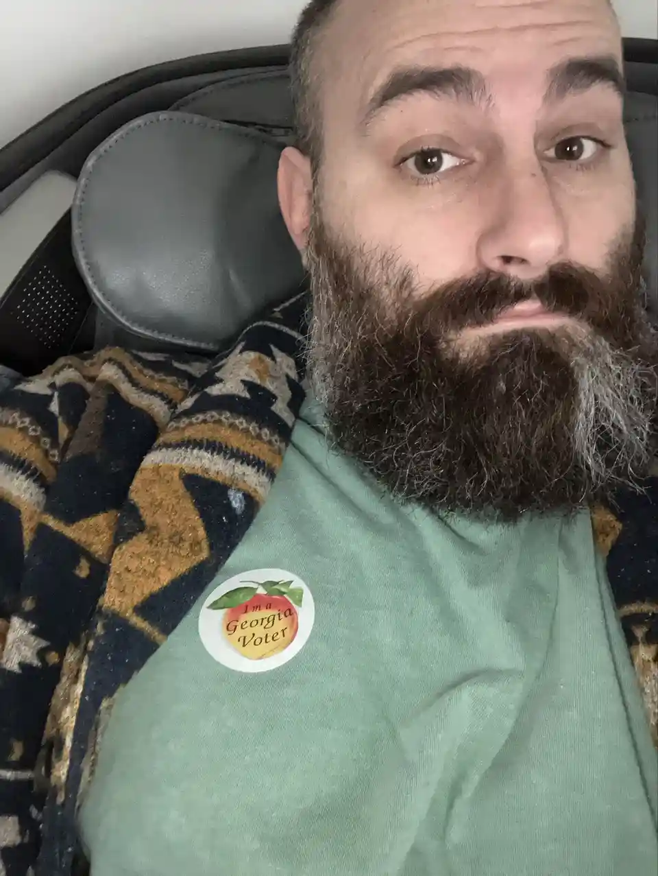 A man with a full beard and mustache, wearing a green t-shirt, sits in a black chair with a patterned blanket draped over…