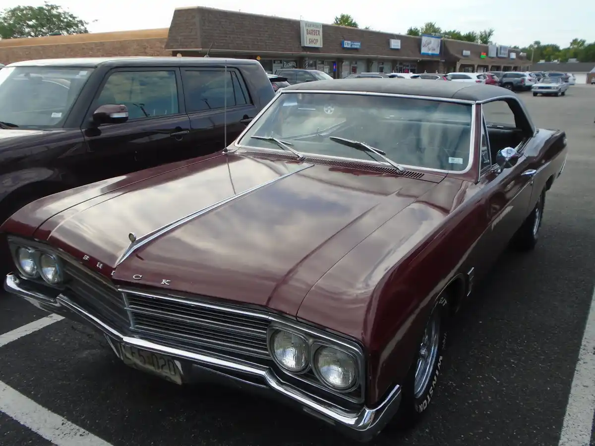 A maroon 1960s Buick with chrome trim and dual headlights is parked in a lot next to a black station wagon, with a 'Hair…
