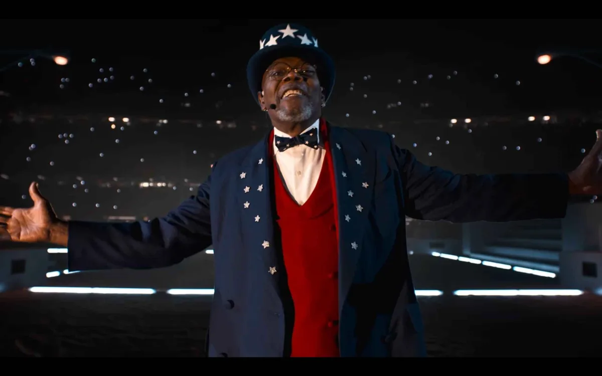 A man dressed as Uncle Sam, wearing a star-adorned top hat, blue jacket, red vest, and bow tie, stands with arms…