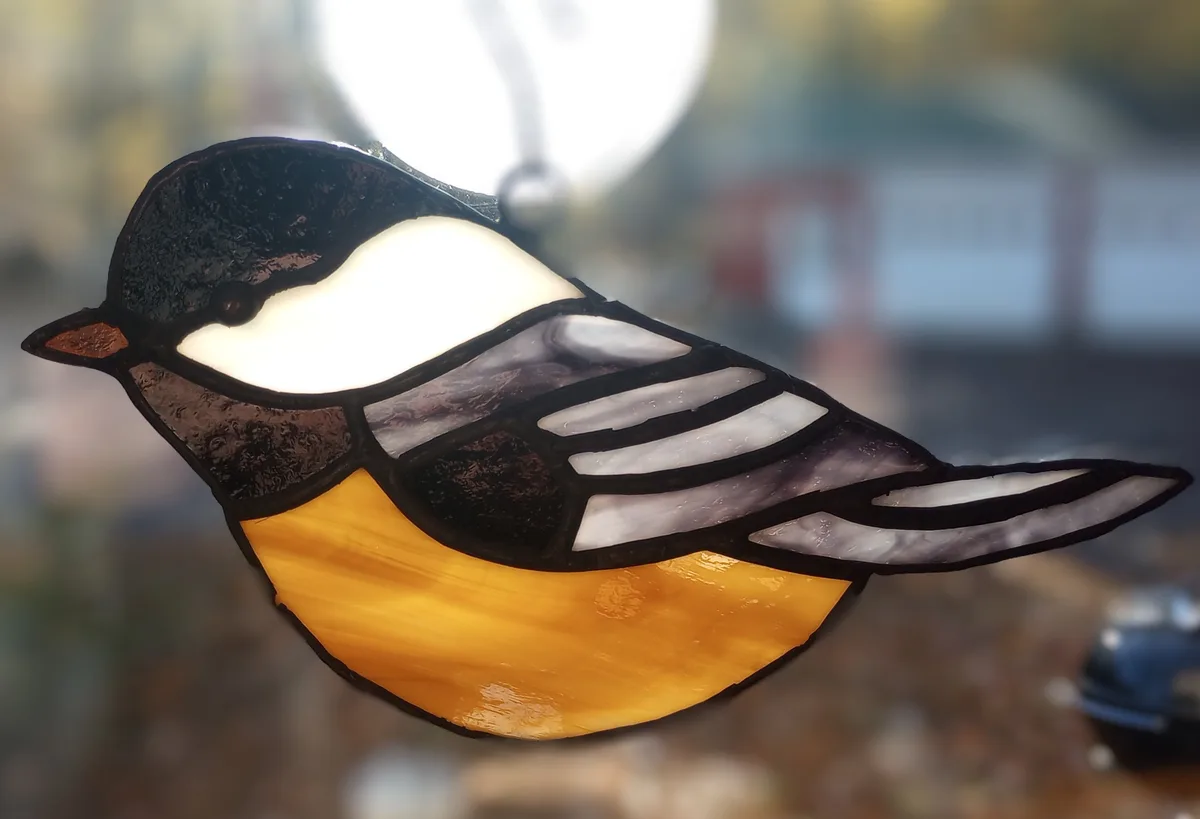 A stained glass suncatcher shaped like a bird with a black head, white cheek, gray and white wings, and a yellow belly…