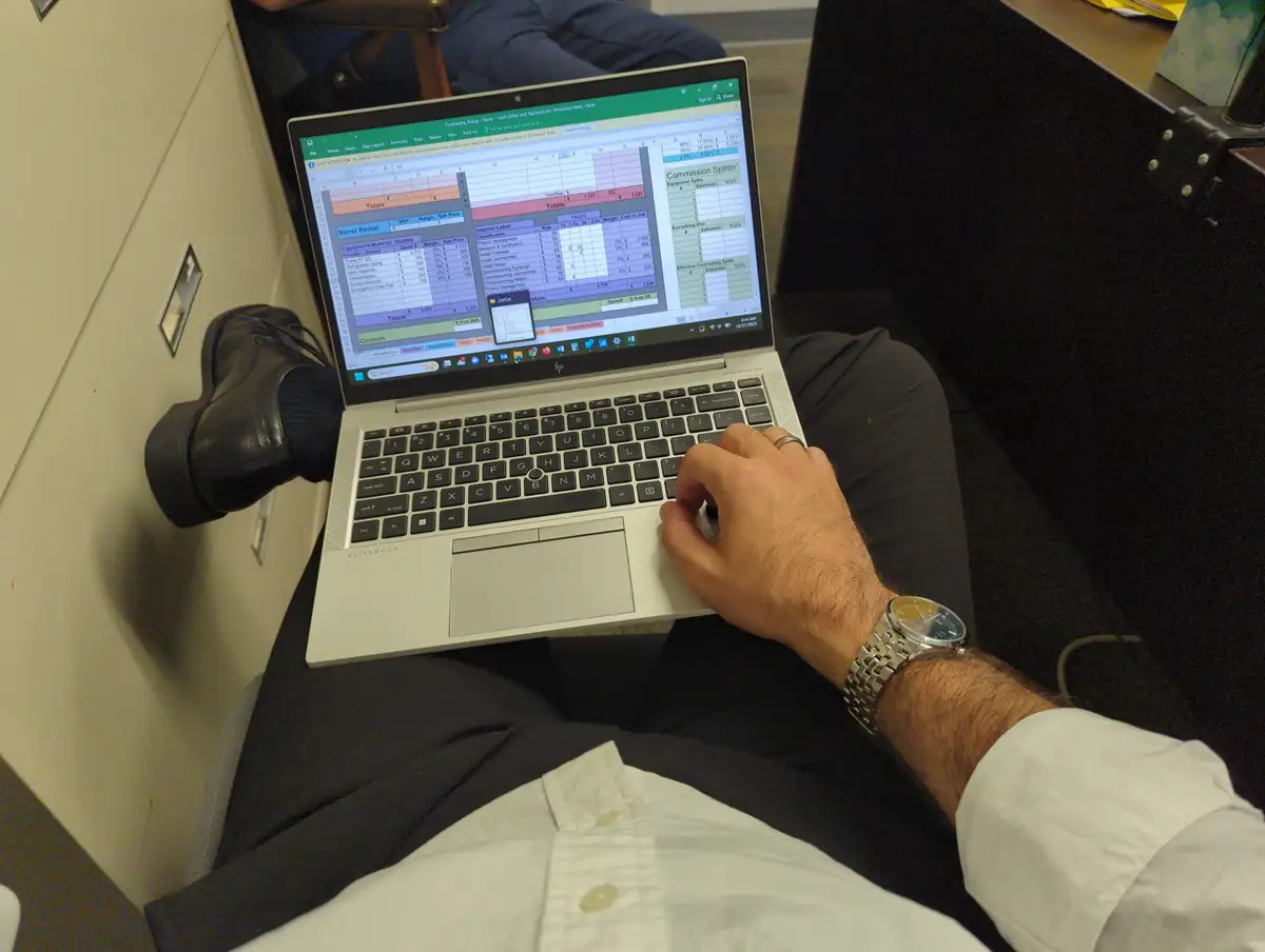 A person is sitting with a laptop on their lap, actively working on a complex Excel spreadsheet filled with financial data…
