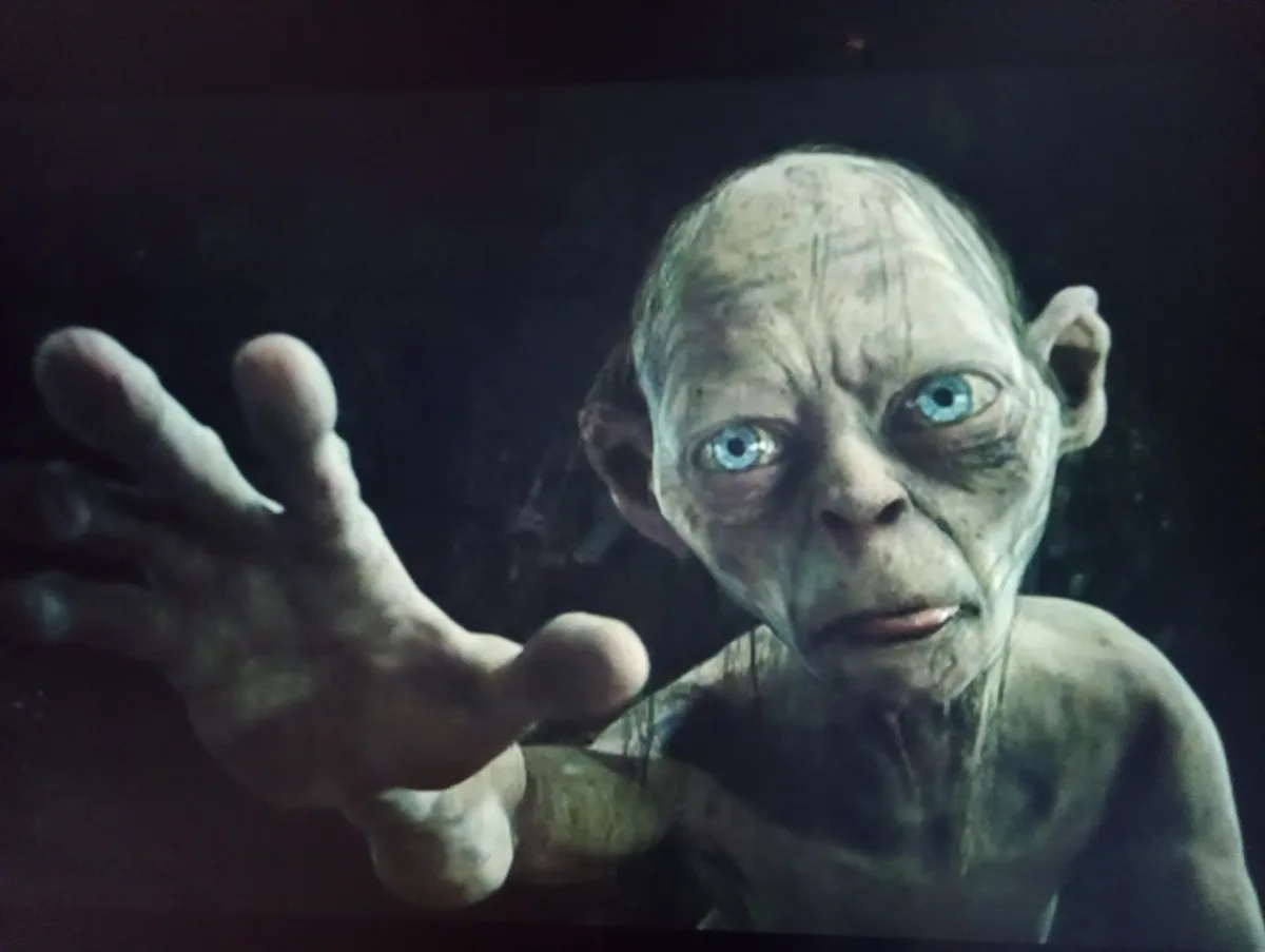 A close-up of the character Gollum from 'The Lord of the Rings' films, with pale, wrinkled skin, large blue eyes, and a hand…