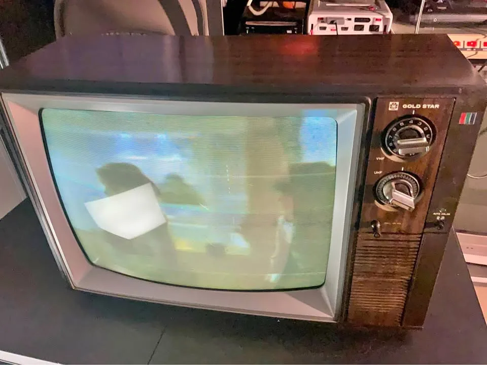 An old, boxy Gold Star CRT television with wooden casing and physical dials sits on a desk, its screen showing static and…