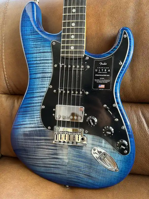 A blue Fender American Ultra Stratocaster HSS electric guitar with a flame maple top and black pickguard rests on a brown…