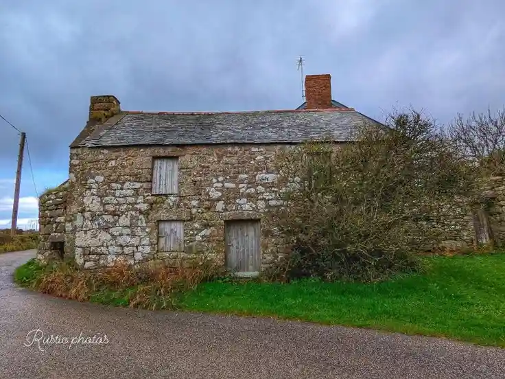 An old, weathered stone cottage with boarded-up windows and a moss-covered roof sits beside a gravel road under a cloudy…