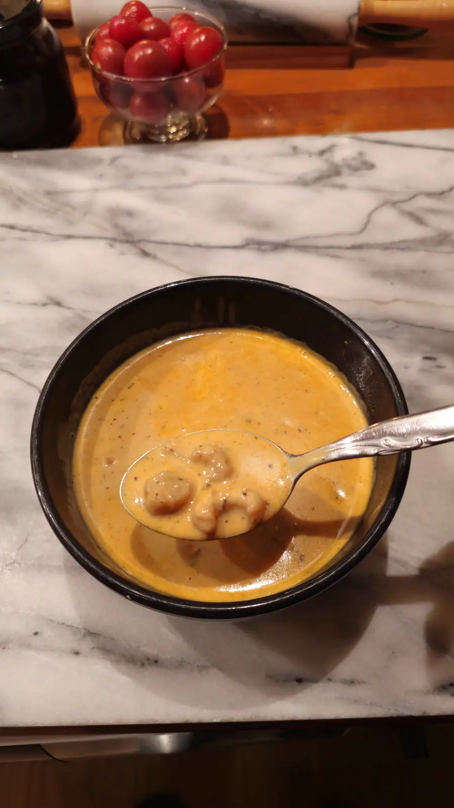 A close-up photo shows a black bowl filled with creamy orange soup on a white marble countertop, with a silver spoon lifting…