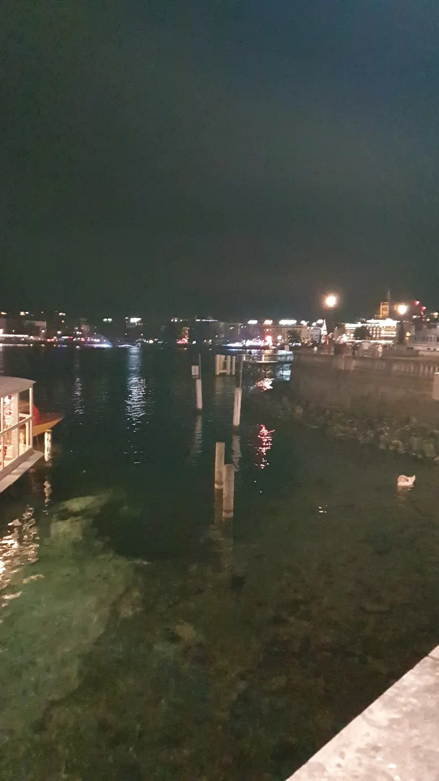 A nighttime photo of a calm waterfront with city lights reflecting on the water, a small white swan swimming alone in the…