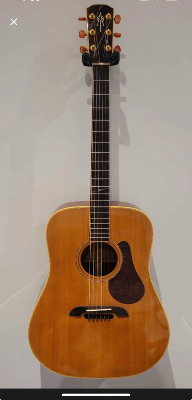 A full-frontal photo of a light-wood acoustic guitar with a black pickguard and headstock logo, standing upright against a…