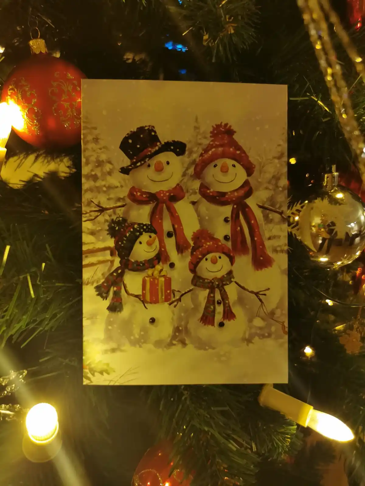 A Christmas card featuring a cheerful family of four snowmen wearing hats and scarves, one holding a gift, displayed against…