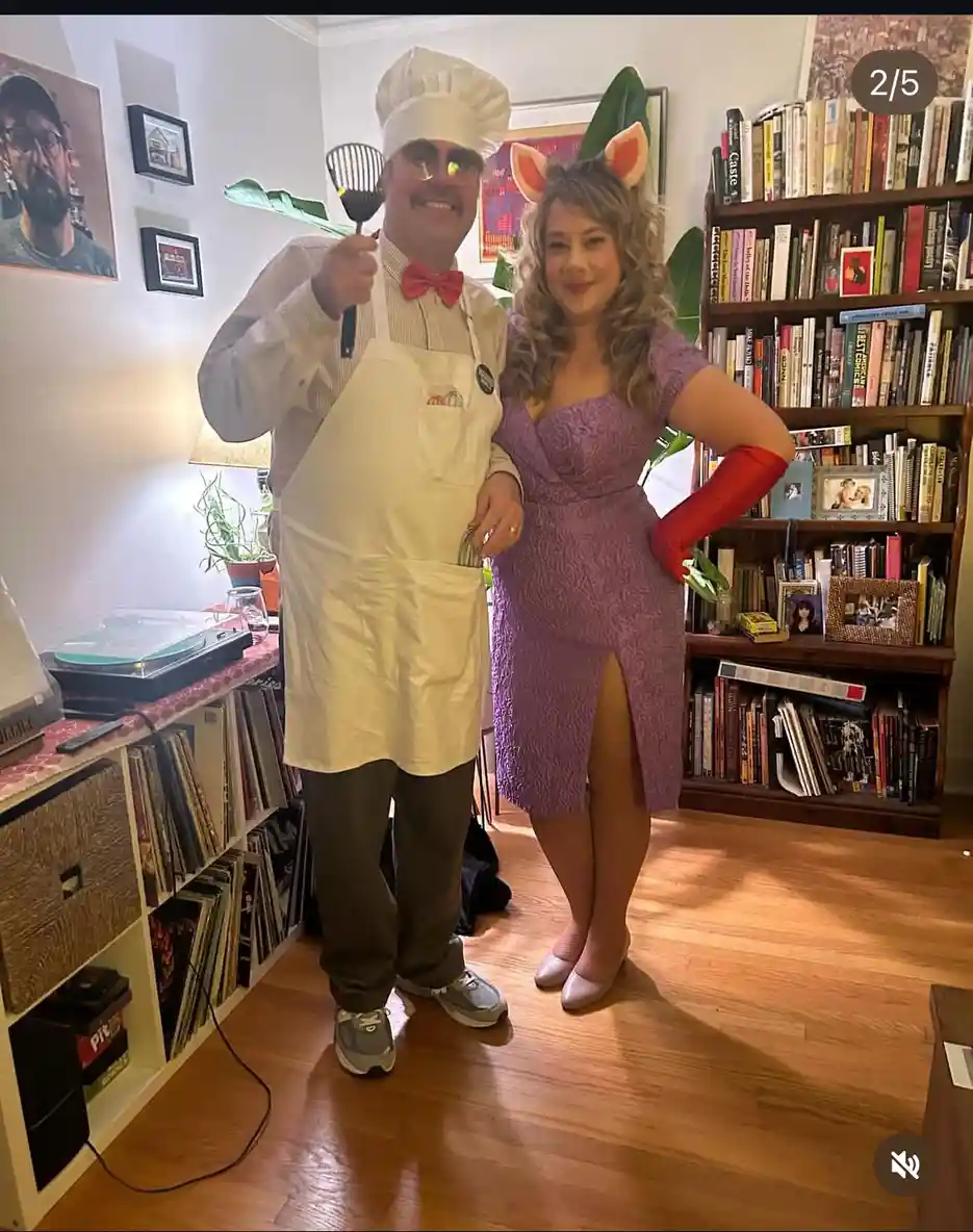 A man dressed as a chef in a white hat, apron, and red bow tie holds a spatula while standing next to a woman in a purple…