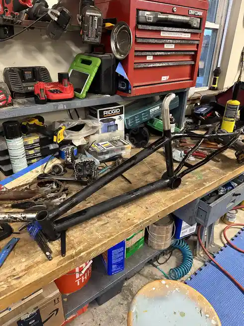 A cluttered workshop workbench holds a black-painted bicycle frame, surrounded by tools, batteries, spray cans, and parts. A…