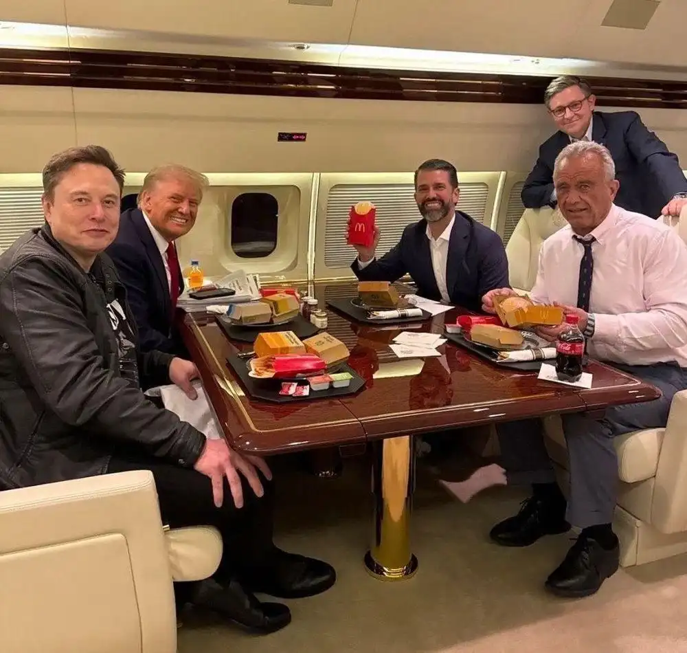 Four men are seated around a polished wooden table inside a private jet, eating McDonald's food from boxes and cups. Elon…