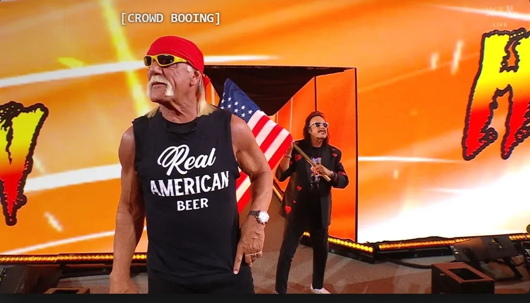A muscular man with a red bandana and sunglasses wears a black tank top that reads 'Real American Beer' while standing on a…