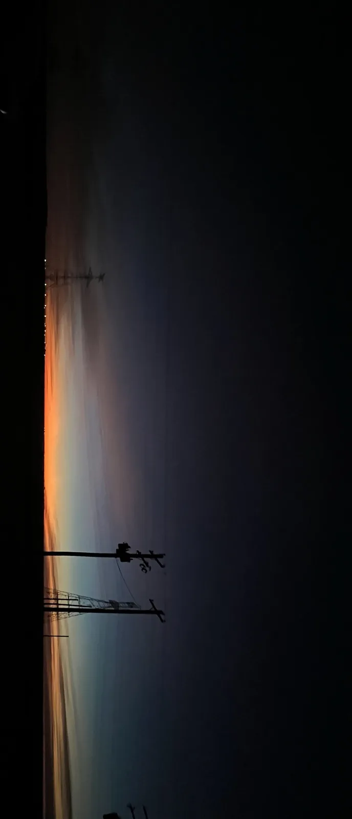 A vertical photo shows a narrow strip of sunset sky glowing orange and blue, framed by blackness on either side, with…