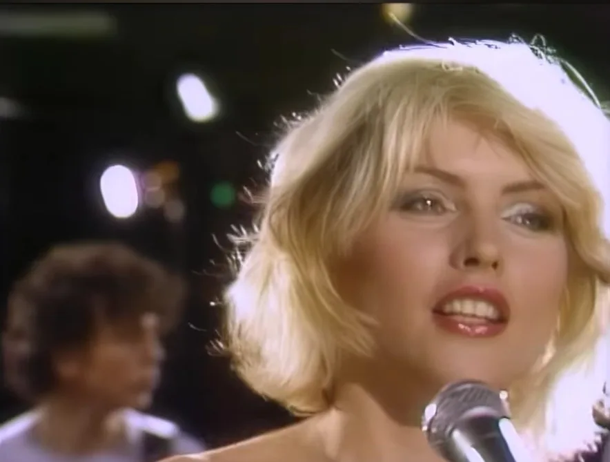 a close-up of a blonde woman singing into a microphone on stage, with a blurred figure behind her. The overlaid text reads…