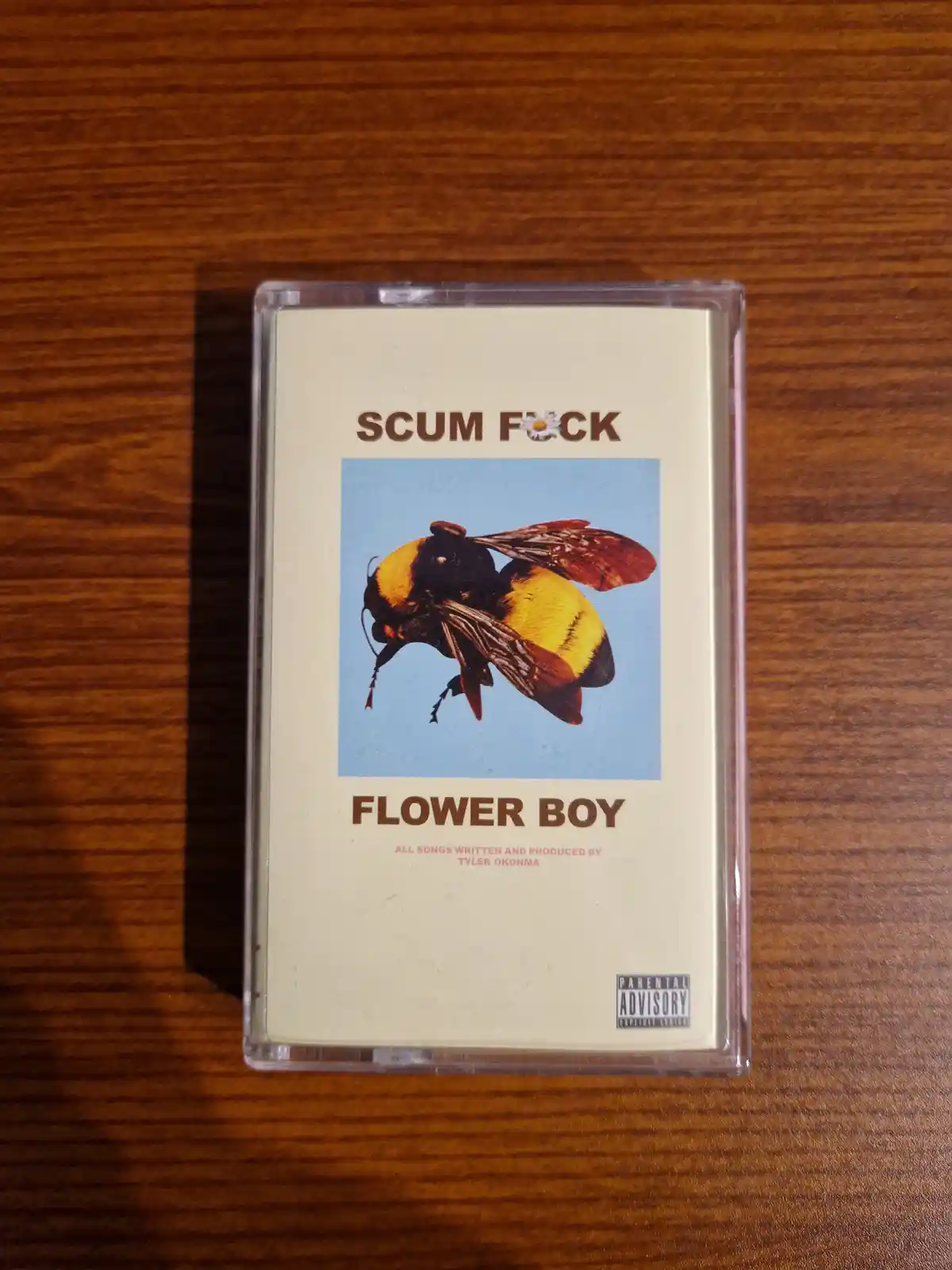 A cassette tape with a beige cover featuring a large bumblebee against a blue sky, the title 'SCUM F*CK' at the top with a…
