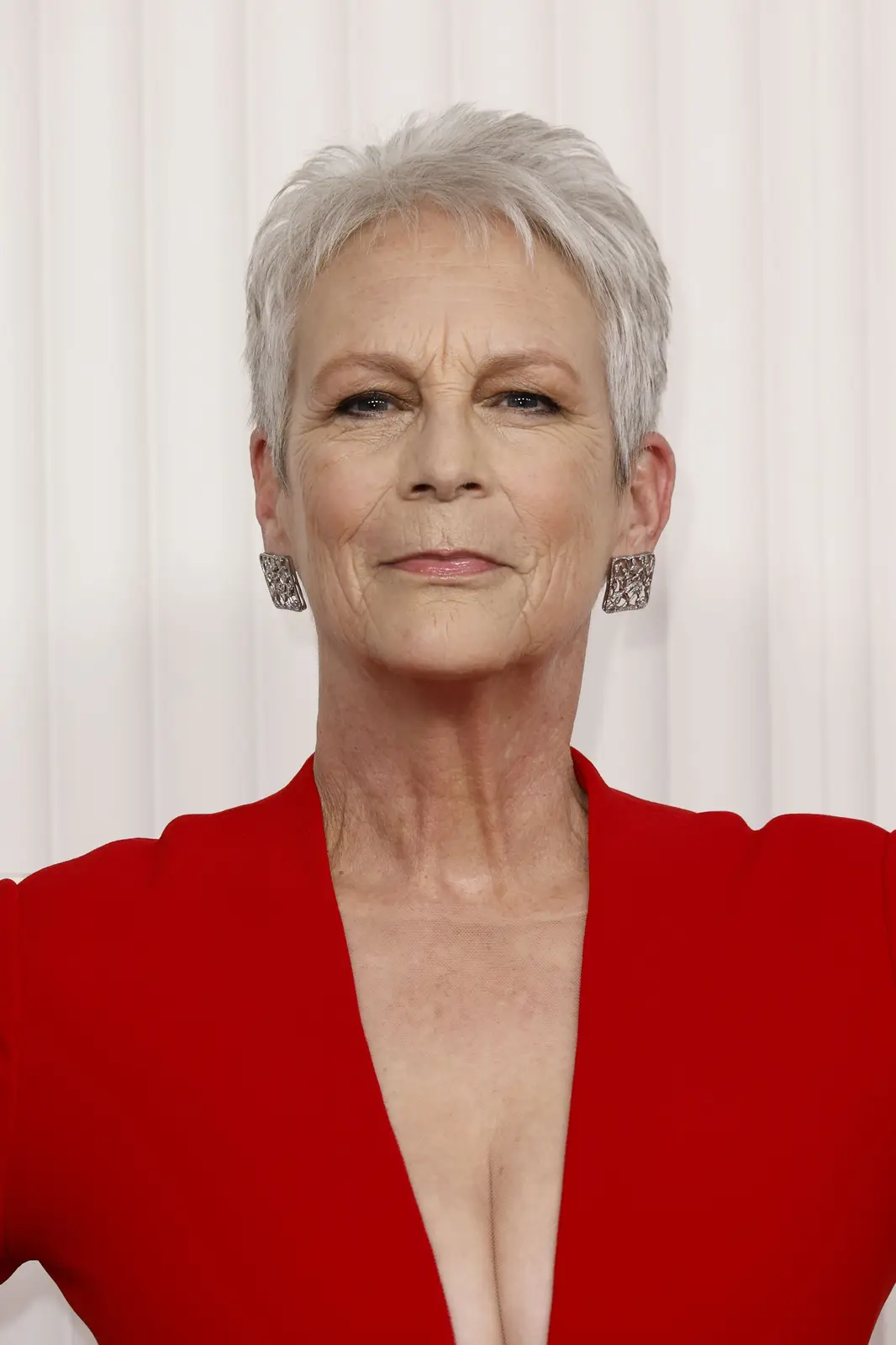 A close-up photo of actress Jamie Lee Curtis, who has short gray hair, is wearing a red dress with a plunging neckline and…
