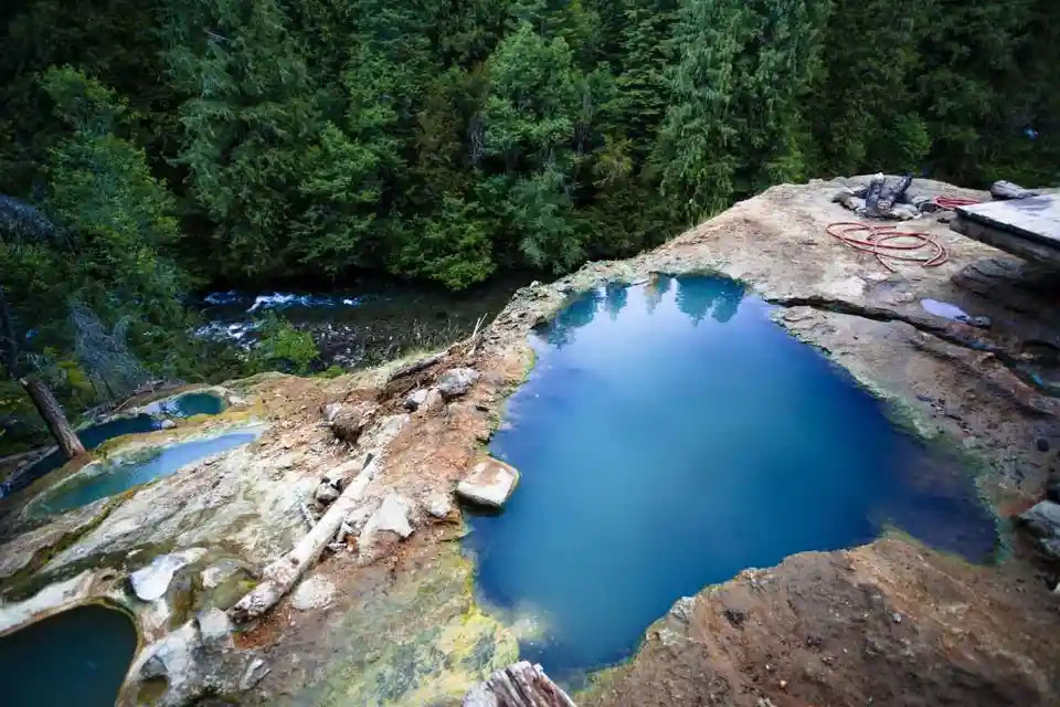 a natural hot spring with vibrant blue water nestled in a forested area, surrounded by earthy, mineral-stained rock…