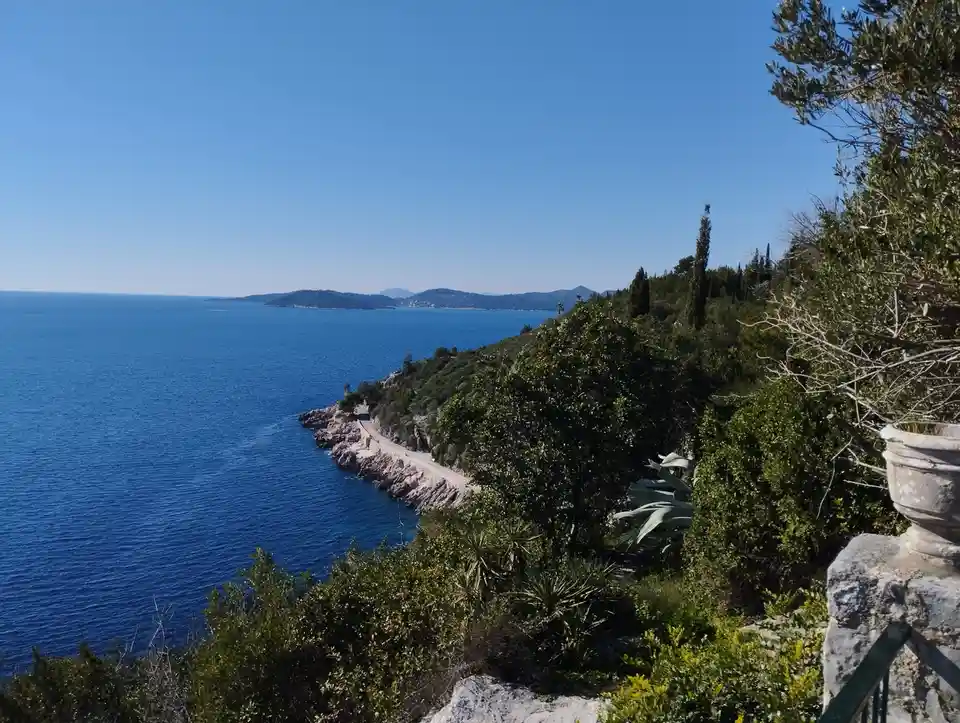 A scenic coastal view shows a winding road along a cliffside overlooking deep blue ocean under a clear sky, with lush…
