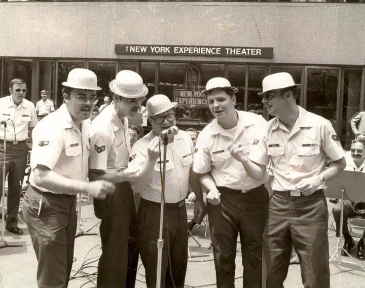 A black-and-white photo shows five men in matching white shirts and white hard hats performing or posing energetically in…
