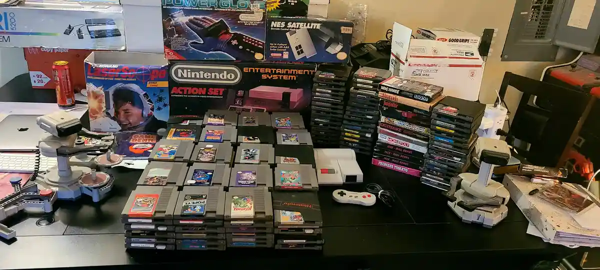 A cluttered desk displays a large collection of vintage Nintendo Entertainment System (NES) items, including numerous game…