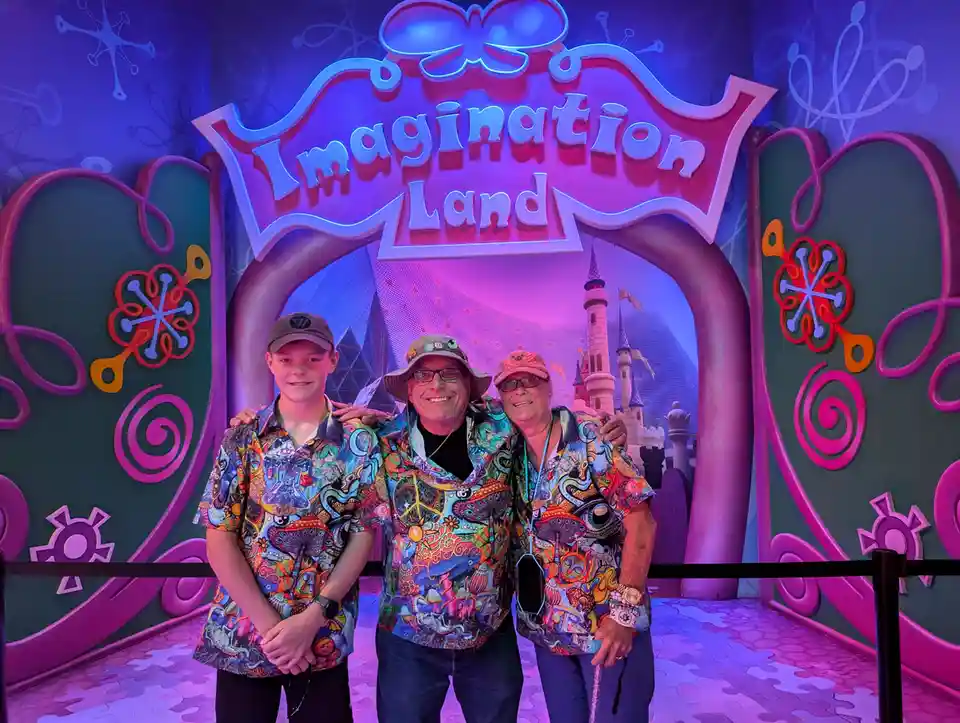 Three people pose together in front of a colorful, whimsical archway labeled 'Imagination Land,' which features cartoonish…