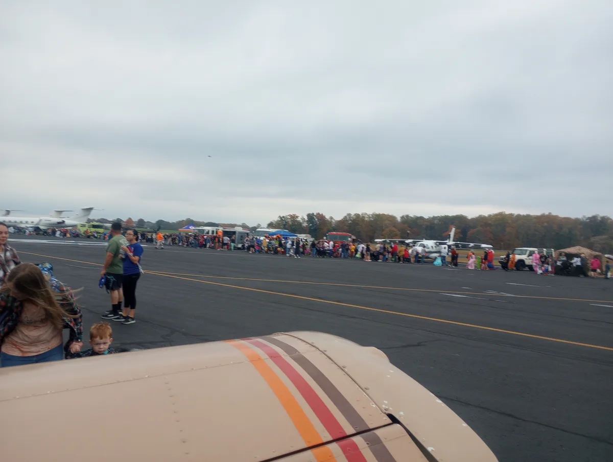 A photo taken from inside a small airplane shows a long line of people on an airport tarmac, many dressed in costumes, with…