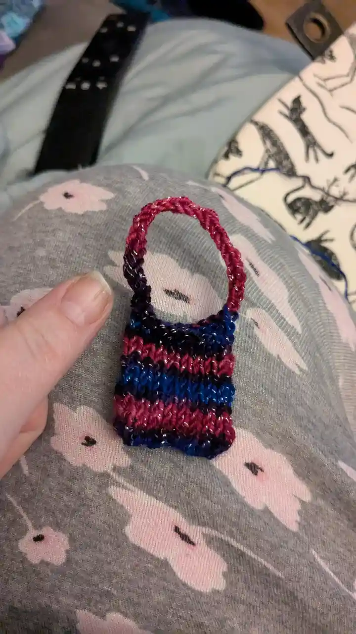 A person's thumb holds a tiny, hand-knitted purse with alternating red and blue stripes and a red handle, resting on…