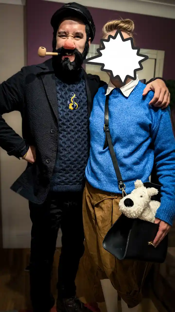 A man dressed as a pirate with a fake beard, red nose, and pipe poses with a woman whose face is obscured by a white…