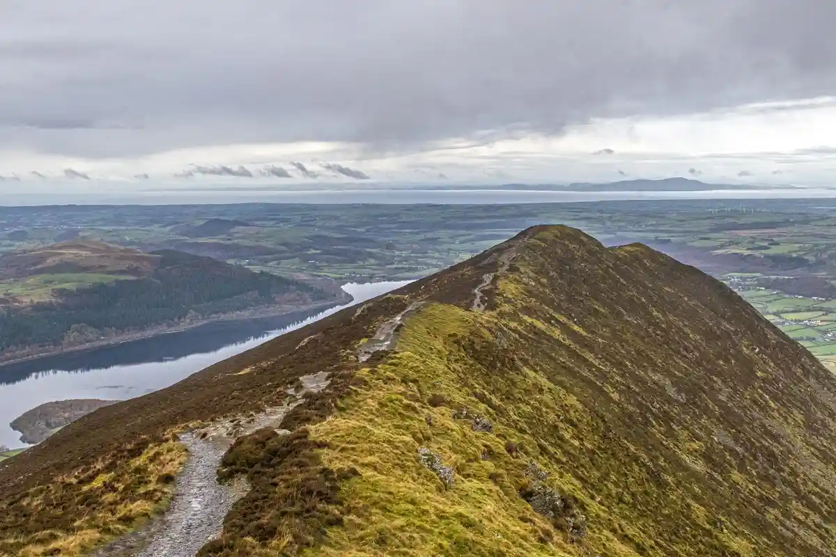 A scenic view from a narrow, grassy mountain ridge with a winding dirt path, overlooking a valley with a lake and distant…