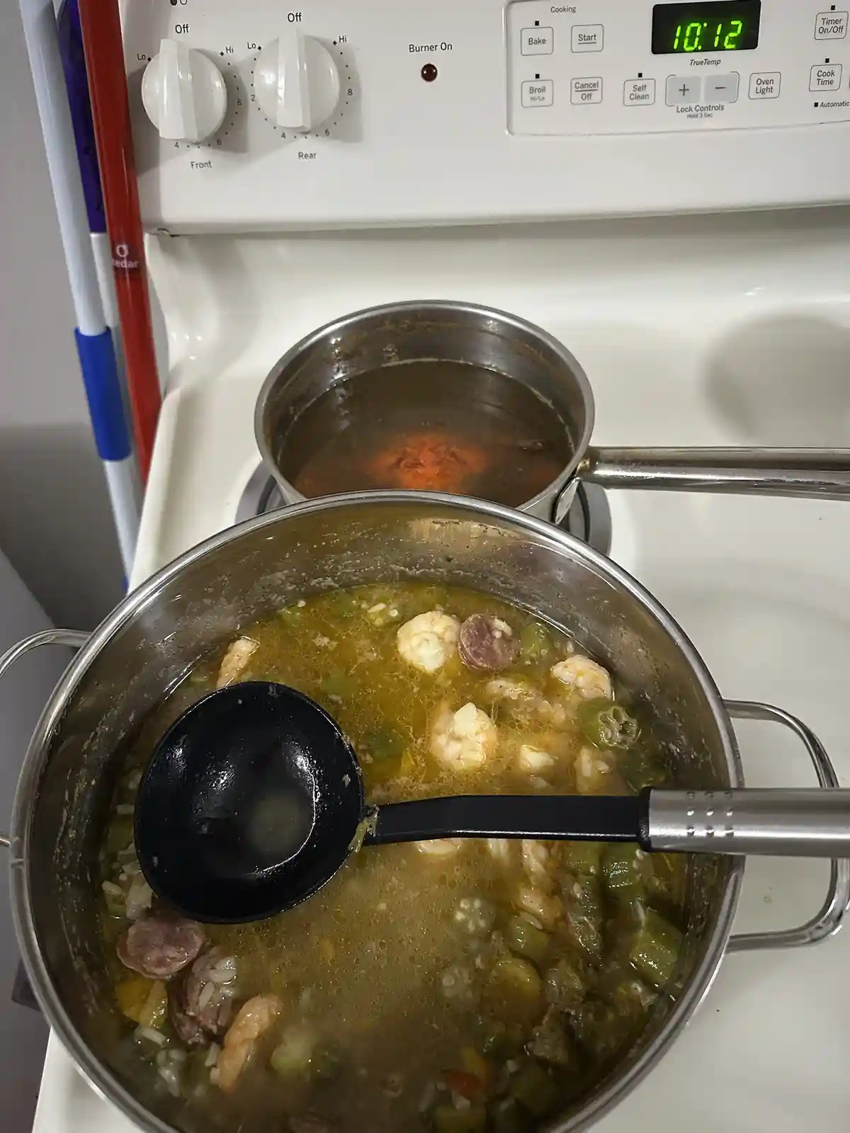 A close-up photo shows two pots on a white electric stove. The front pot contains bubbling jambalaya with shrimp, sausage…