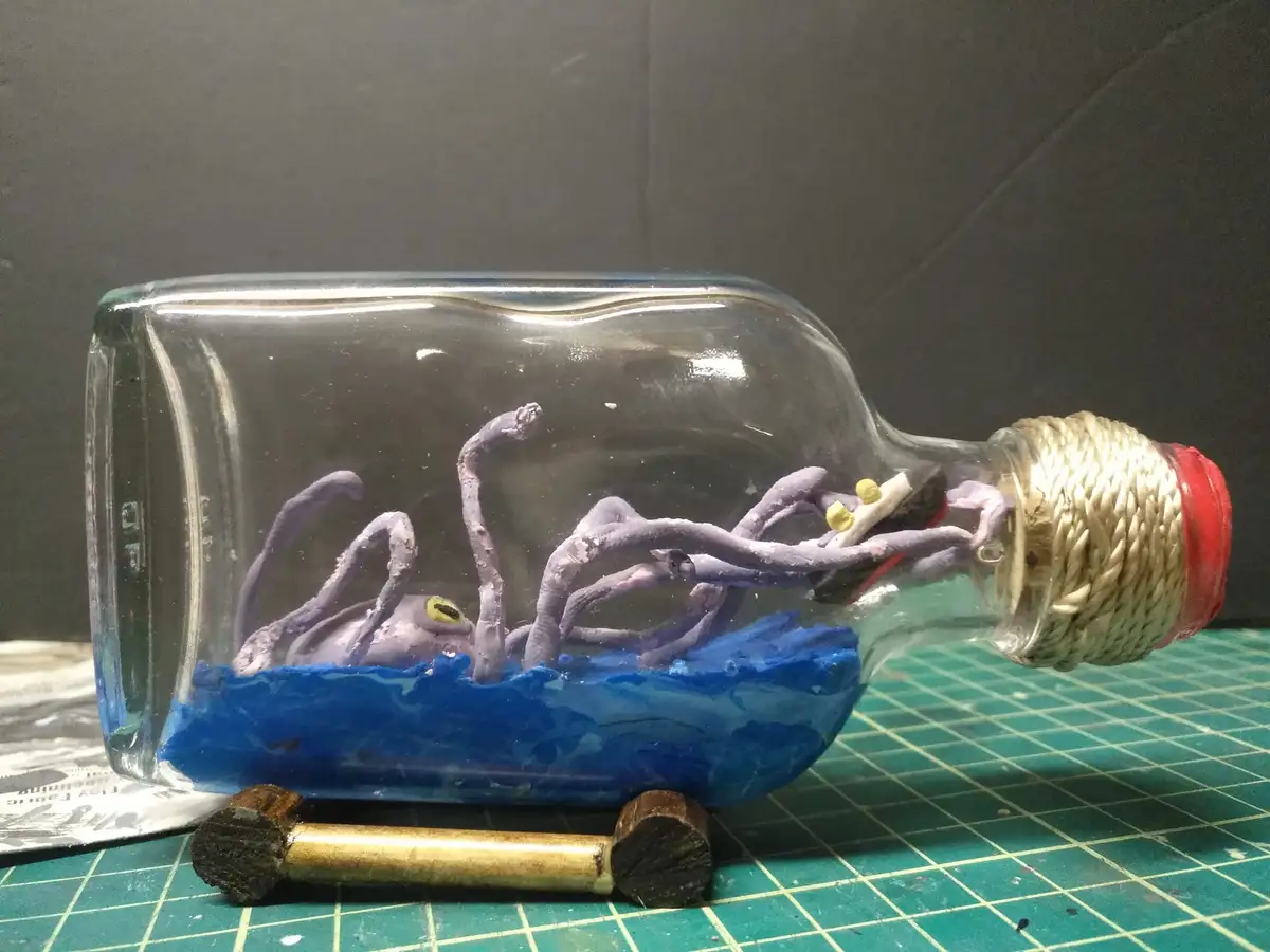 A handmade diorama inside a clear glass bottle shows a purple clay octopus with yellow eyes, partially submerged in blue…