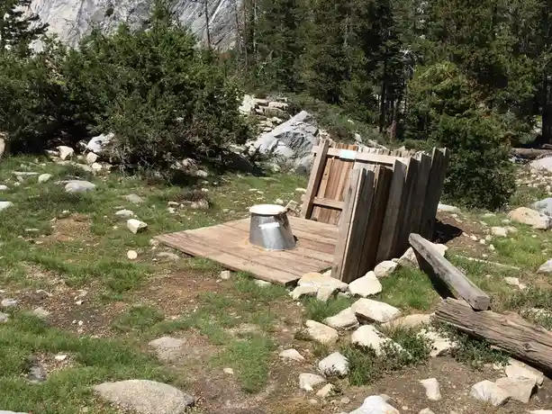 An outdoor wooden structure resembling a rustic outhouse in a mountainous, forested area with rocks and grass, featuring a…