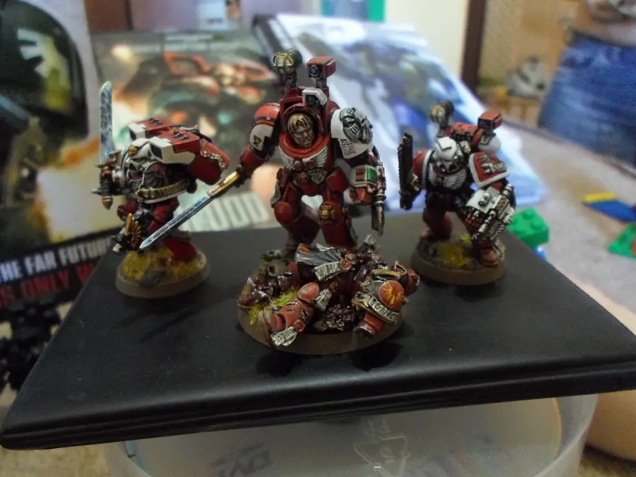 Three painted red-and-white armored miniature figures stand on a black display stand, one fallen at their feet. Behind them…