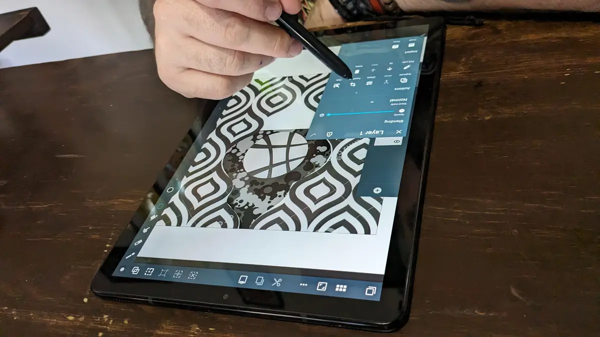 A person's hand holds a stylus over a tablet displaying a digital drawing app with a black-and-white geometric pattern. The…