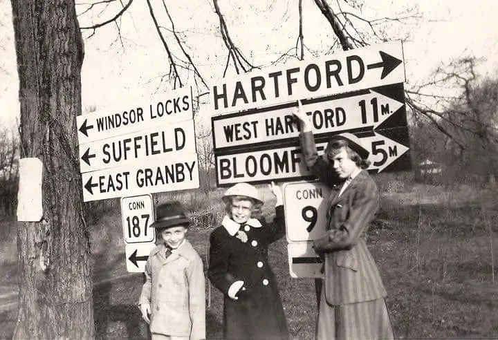 A vintage black-and-white photo shows three young girls posing beside a cluster of directional road signs. One girl holds up…