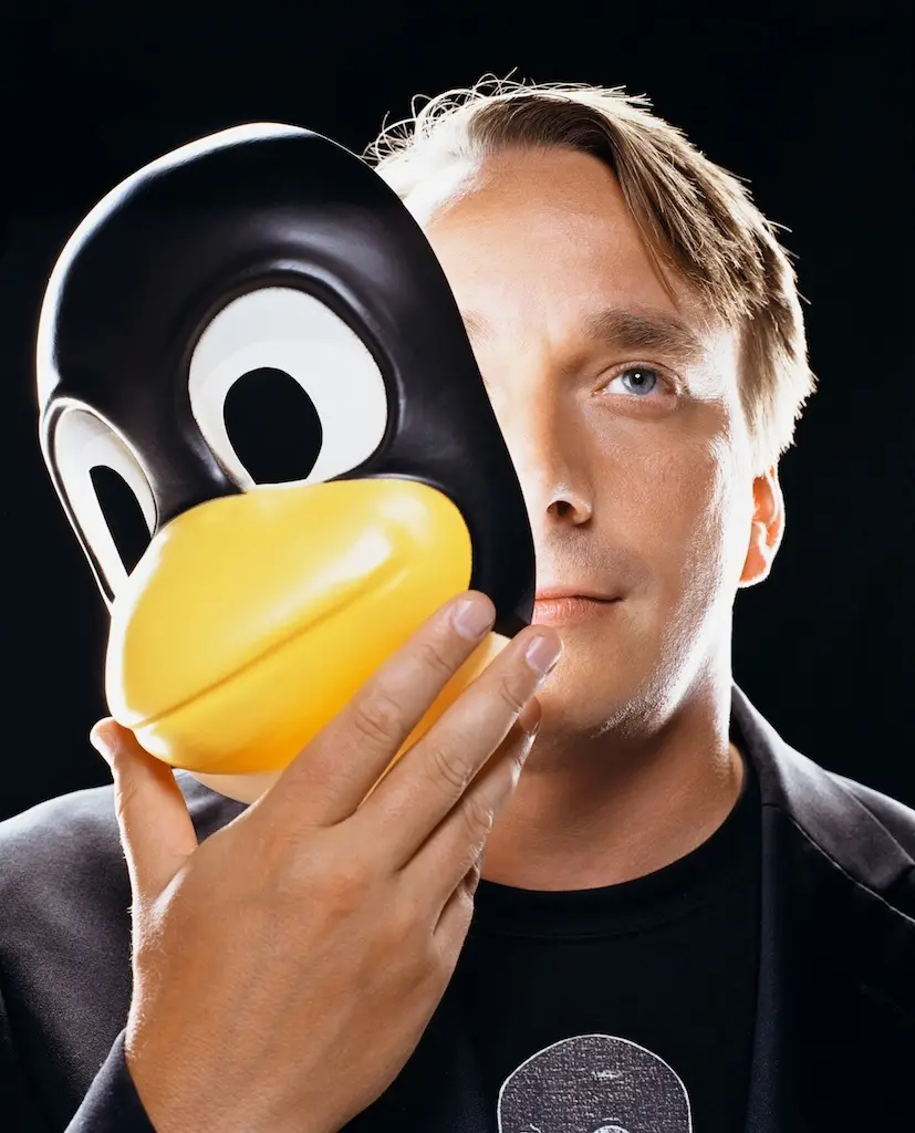 A man with light brown hair and blue eyes holds a large, glossy black-and-yellow penguin mask in front of his face, covering…