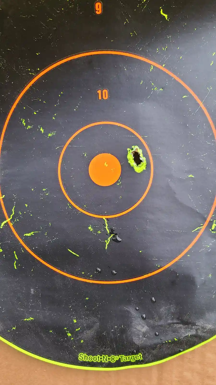 A close-up of a black shooting target with orange scoring rings, labeled '10' and '9'. A large, ragged hole is in the center…