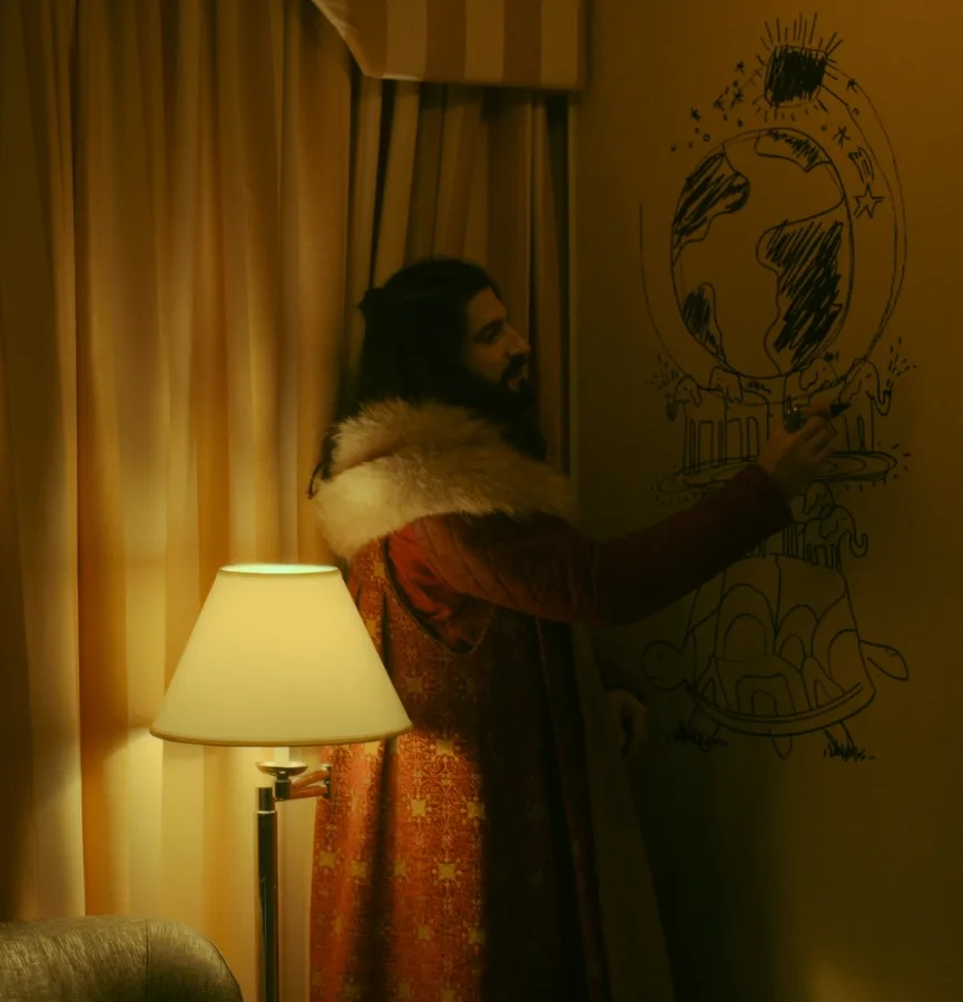 A man in a red robe with a fur collar stands in a warmly lit room, drawing a crude sketch of the Earth on a wall. The…