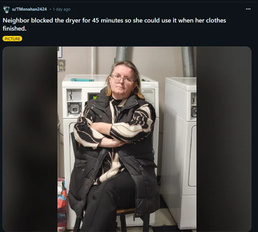 A woman with glasses and a black puffer vest sits on a chair in a laundromat, arms crossed, looking annoyed. Behind her is a…