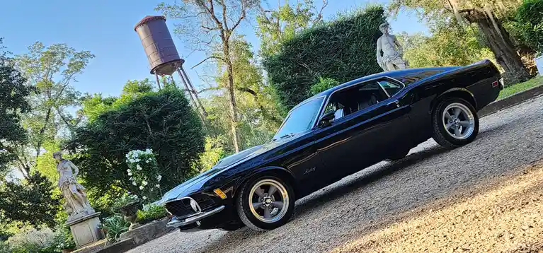 A glossy black classic muscle car, likely a Ford Mustang, is parked on a gravel driveway surrounded by lush greenery…