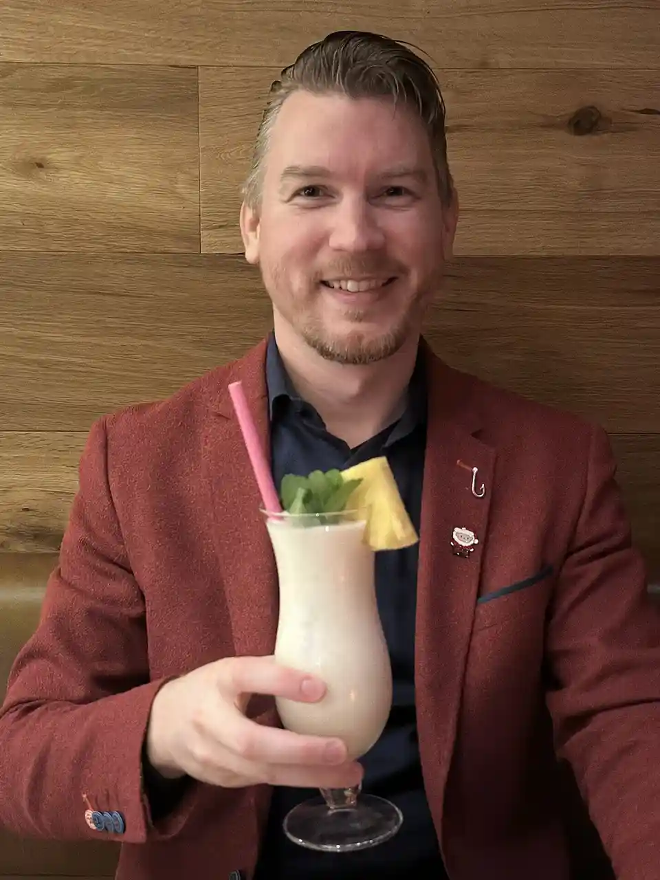 A smiling man in a red blazer holds a creamy tropical drink garnished with a pineapple slice and mint, with a pink straw. He…