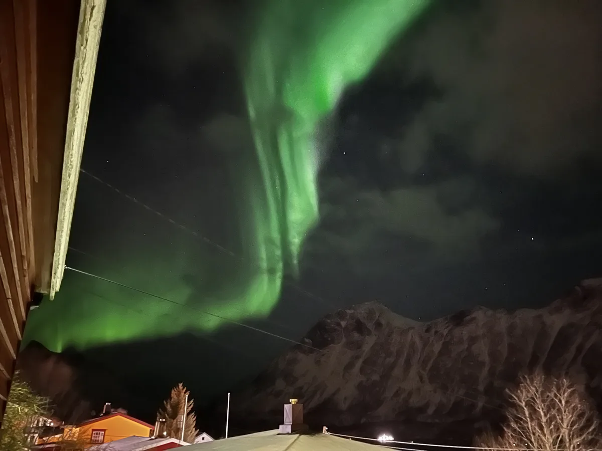 A nighttime photo shows vibrant green aurora borealis dancing across a starry sky above snow-dusted mountains and rooftops…