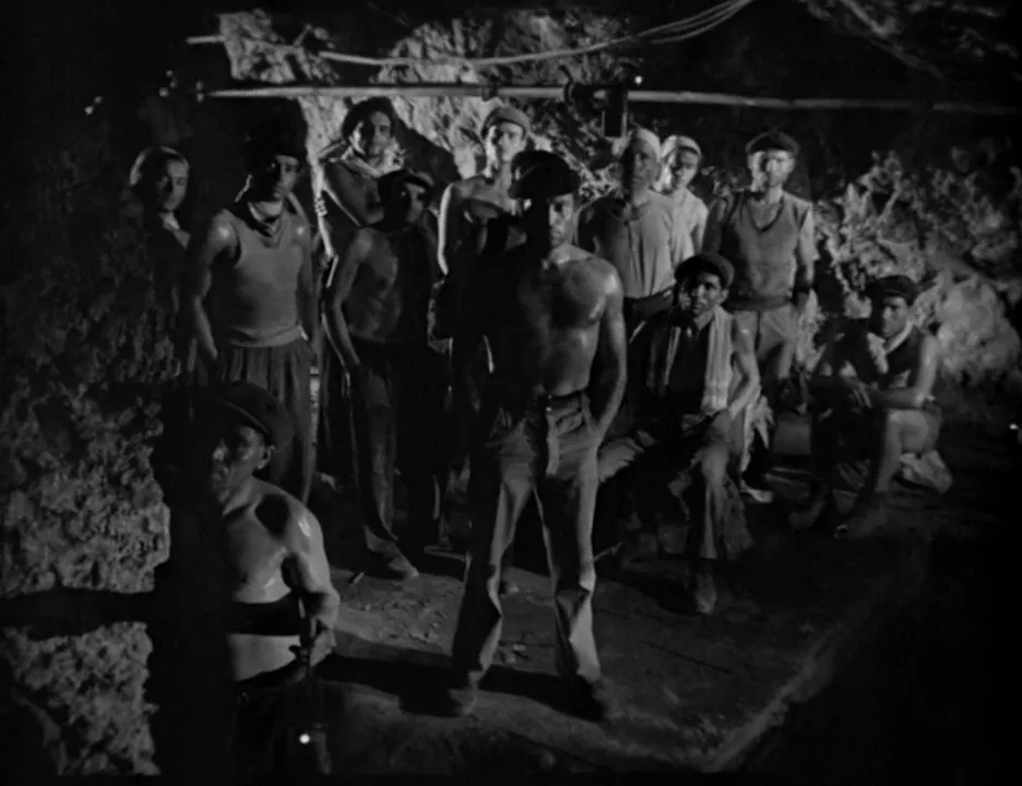 A black-and-white still from a film shows a group of shirtless, sweaty men in a dimly lit, rocky underground setting…