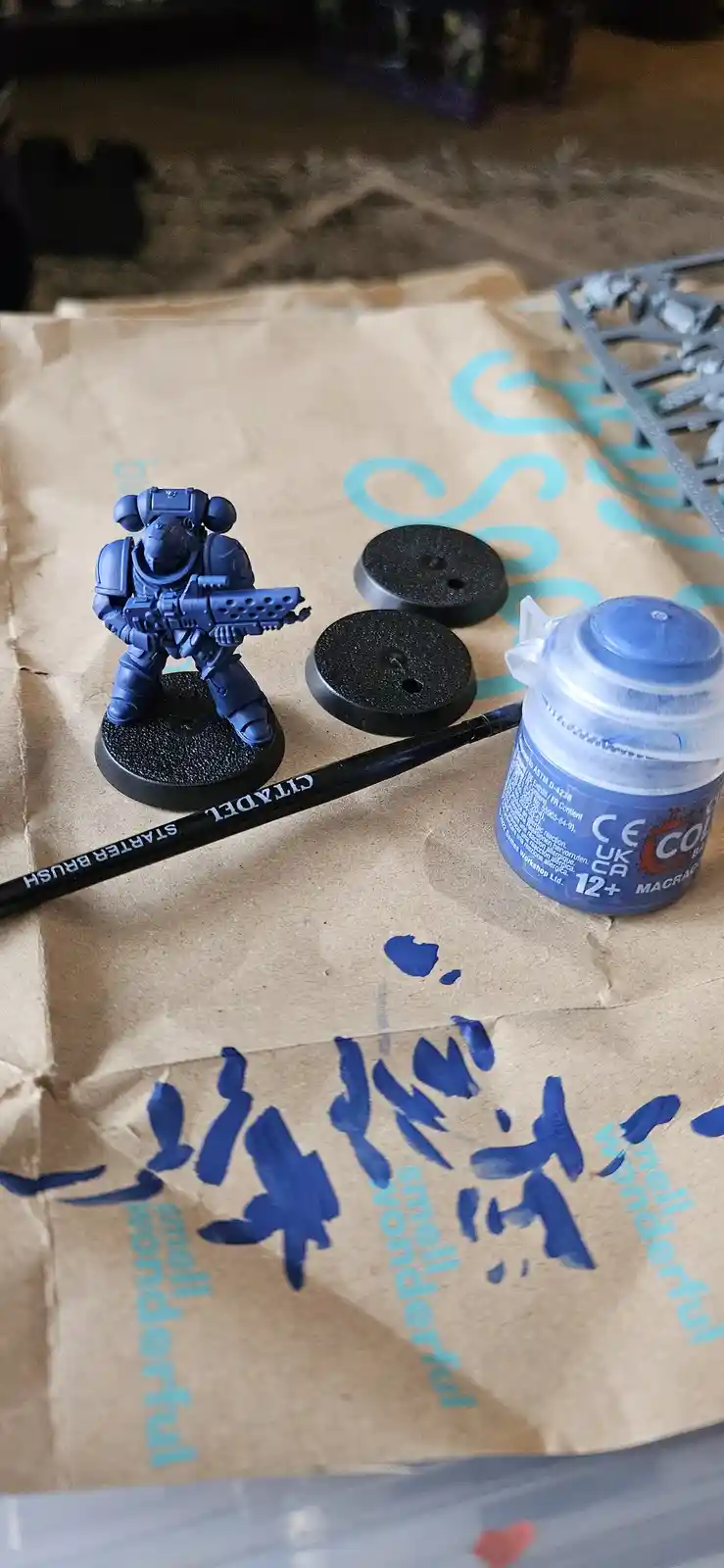 A blue-painted miniature warrior figurine stands on a black base next to two spare bases, a Citadel starter brush, and a…