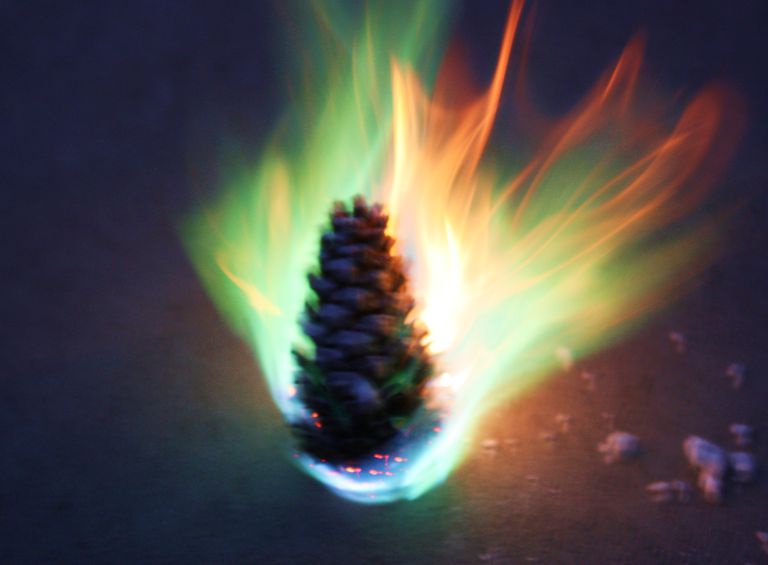 A pinecone is engulfed in vibrant, blurred flames that radiate outward in streaks of orange, yellow, and green against a…