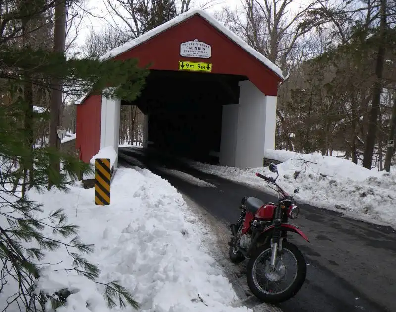 A red motorcycle is parked on a snowy road in front of a red and white covered bridge. A yellow sign above the bridge…